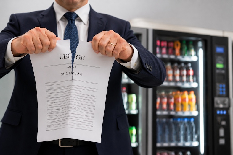 Politico che strappa simbolicamente un documento legislativo sulla sugar tax davanti a distributori automatici di bevande